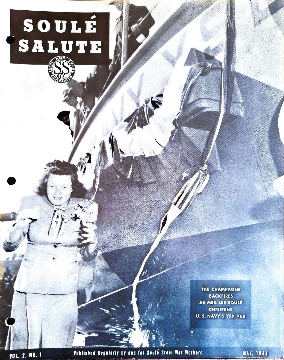 A woman in a suit stands on a dock while a ship is christened with a broken bottle.